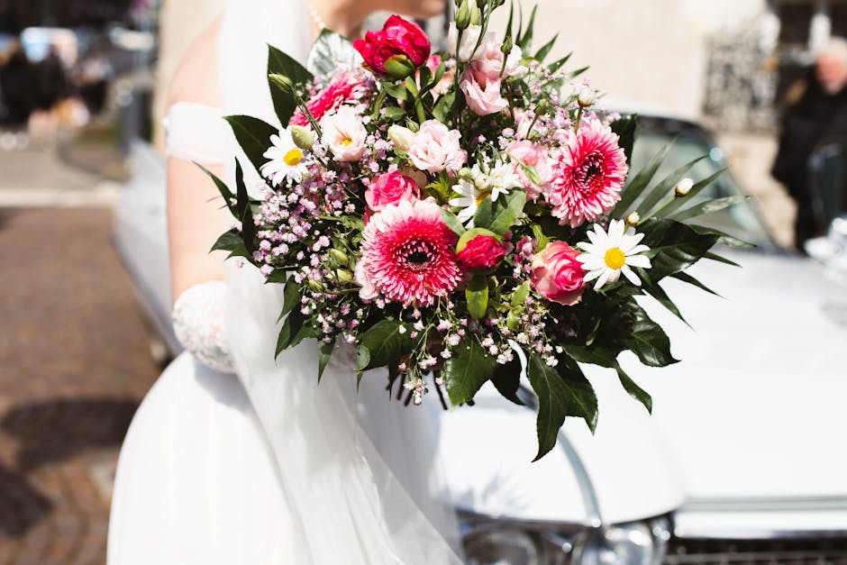 A vibrant bouquet of fresh flowers held by a bride in a white wedding gown, featuring pink and white blooms including gerbera daisies, roses, and baby's breath, with lush green leaves. The arrangement