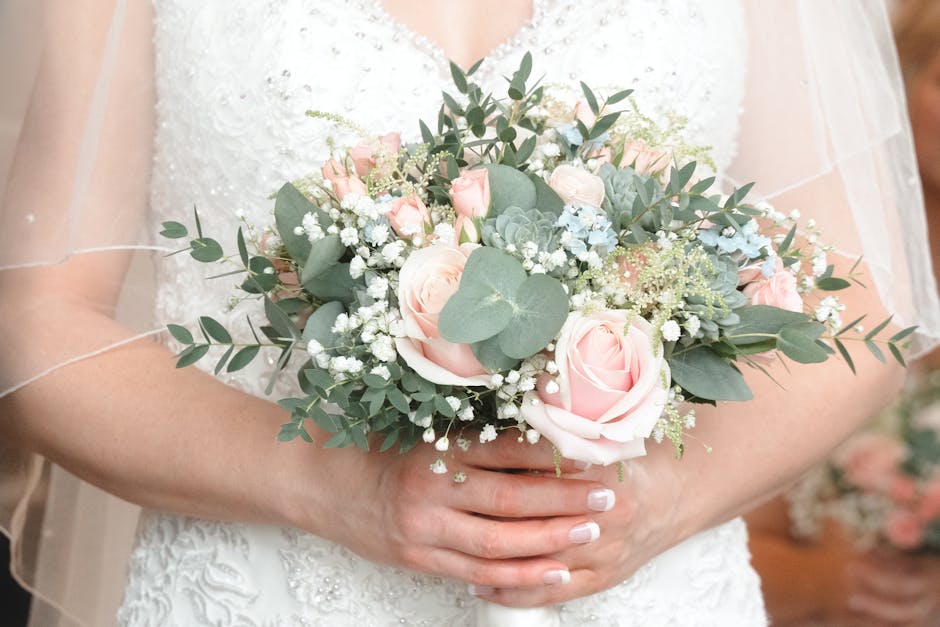 A close-up of a bride holding a floral bouquet arrangement featuring soft pink roses, delicate white baby's breath, and green eucalyptus leaves. The bouquet is accentuated with a white ribbon wrapping