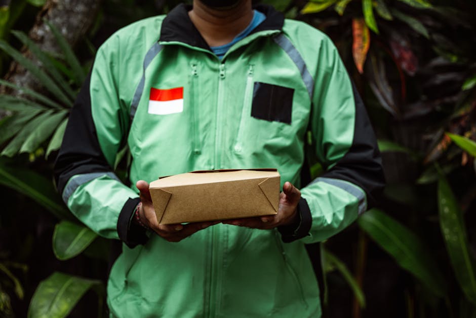 A delivery person wearing a green uniform with black and neon accents, featuring a red and white flag patch on the chest, is holding a rectangular brown cardboard parcel with both hands. The backgroun