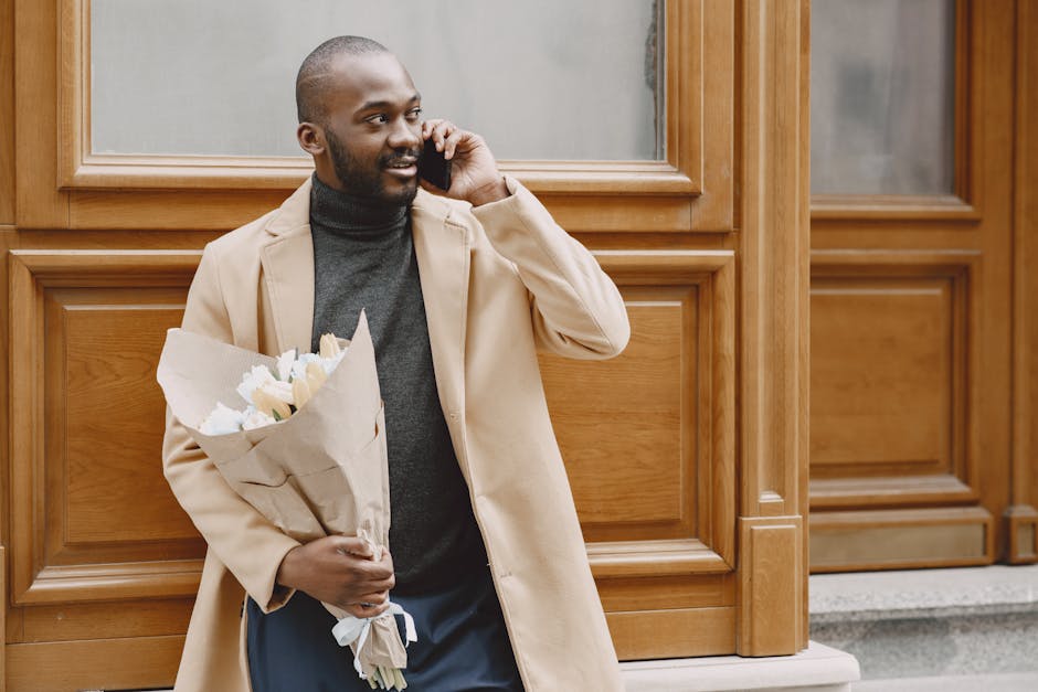 A man with a dark complexion, short hair, and a beard is standing outside in front of wooden paneling. He is holding a bouquet of fresh white tulips wrapped in brown craft paper and tied with a white