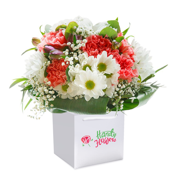 An exquisite, heartwarming bouquet is expertly displayed upright in a modern, white square gift bag-its clean lines and sturdy ribbon handles reflecting a sense of contemporary elegance often found in Bexleyheath's local boutiques. The arrangement blossoms with peach and coral-hued carnations, their lavishly ruffled petals layered thick, creating a tactile sensation of plush abundance. Peppered between, white daisy-like chrysanthemums with vivid lime-green centres add a pop of revitalising energy, reminiscent of fresh blooms seen along the Green Chain Walk early in the season. Wispy sprigs of baby's breath-each tiny petal like a miniature cloud-lend an airy and innocent accent to the more substantial forms. Resting gently on the left side, purple-edged buds, likely alstroemeria, suggest new beginnings and hint at unfolding stories. Encircling the flowers, a collar of deep, shiny green leaves provides balance, structure, and a rich visual anchor, evoking the peaceful canopy of Martens Grove Park. The overall shape of the bouquet is full-bodied and round, a symbol of abundance and care. Against a pristine white background, the vivid palette and thoughtful composition stand out, making this arrangement perfect for occasions from birthdays to anniversaries, or as a fresh surprise-available for prompt delivery in and beyond Bexleyheath.