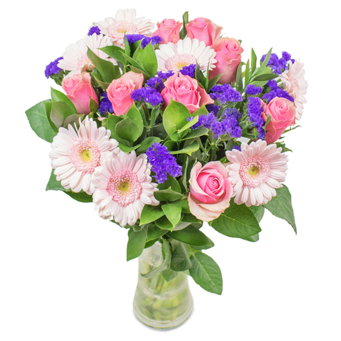 This eye-catching arrangement, elegantly presented in a tall, clear glass vase, invites a sense of freshness reminiscent of a Bexleyheath garden at first light. The bouquet features approximately seven to nine soft and medium pink roses, some with petals just beginning to open in a classic spiral, their colour gently blushing like the clouds above nearby Danson Park. Scattered among them sit five to seven pale pink Gerbera daisies, easily recognized by their large, round faces and pale centres glowing softly like morning dew on Market Place. Deep green foliage creates a lush, abundant foundation, framing the pink blooms while clusters of vibrant purple statice-or perhaps Limonium-are artfully interwoven, adding dynamic texture and rich, jewel-toned accents. The visible stems, submerged in crystal-clear water, highlight the bouquet's vitality, echoing the freshness of a new flower delivery from Flowers Bexleyheath. Ideal for milestone birthdays, heartfelt anniversaries, Mother's Day, or as a sophisticated accent for a welcoming Bexleyheath home or bright office on Albion Road, this arrangement embodies joy, celebration, and authentic affection-each detail thoughtfully curated to reflect the spirit and beauty of the local area.